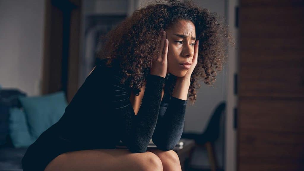 A woman sitting indoors with her hands on her face, looking worried.