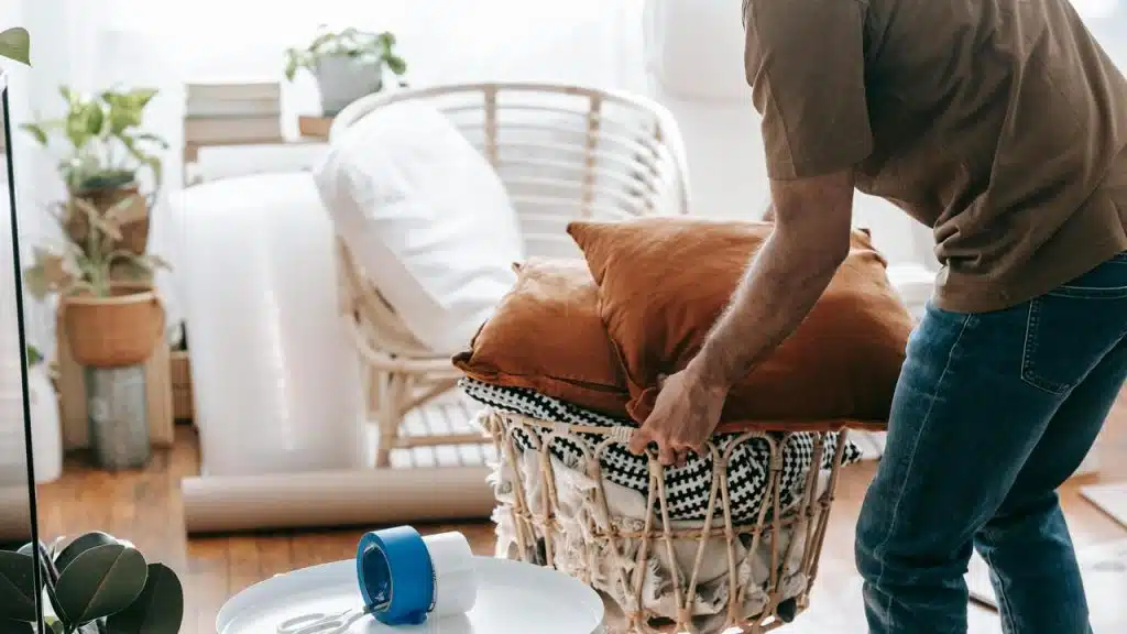 A person packing pillows and blankets into a basket in a cozy room.