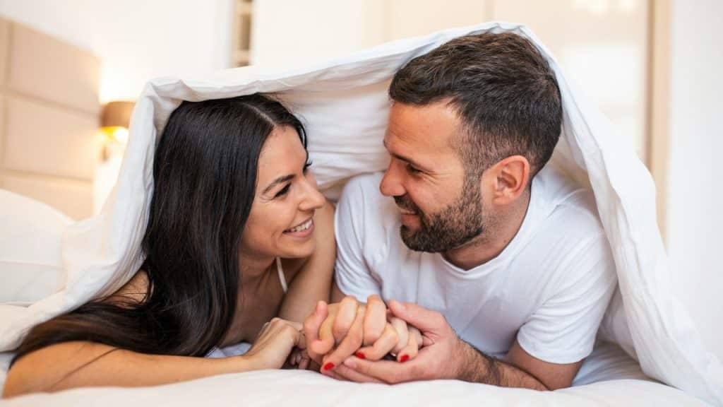A smiling man and woman are lying in bed, holding hands under a white blanket.