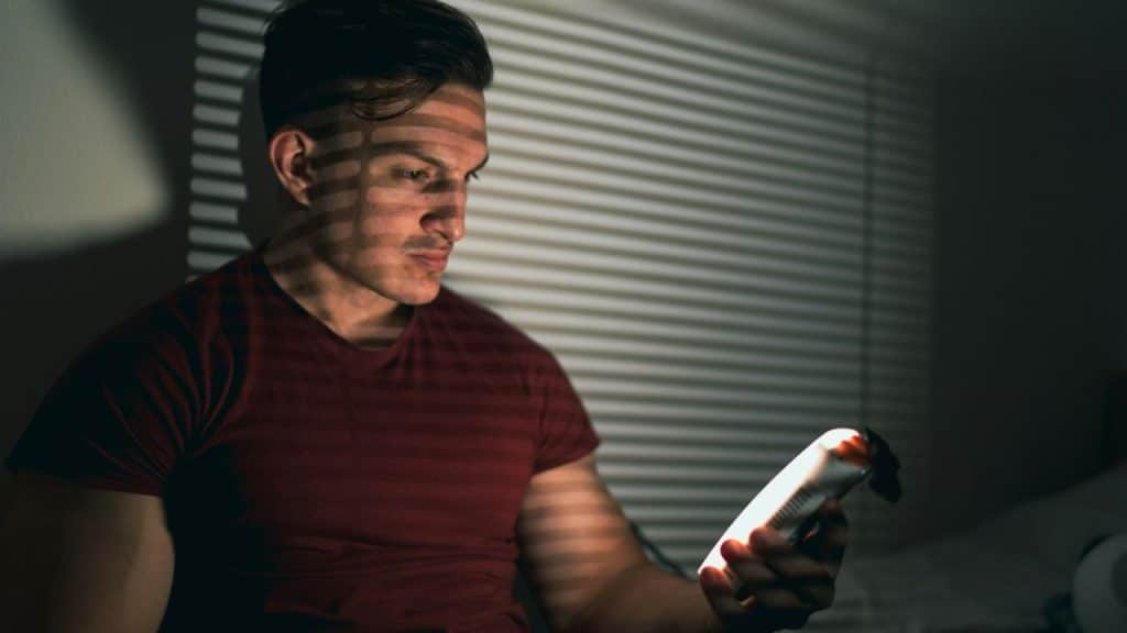 A serious man in a red shirt is looking at a phone in a dark room with window blind shadows on his face.
