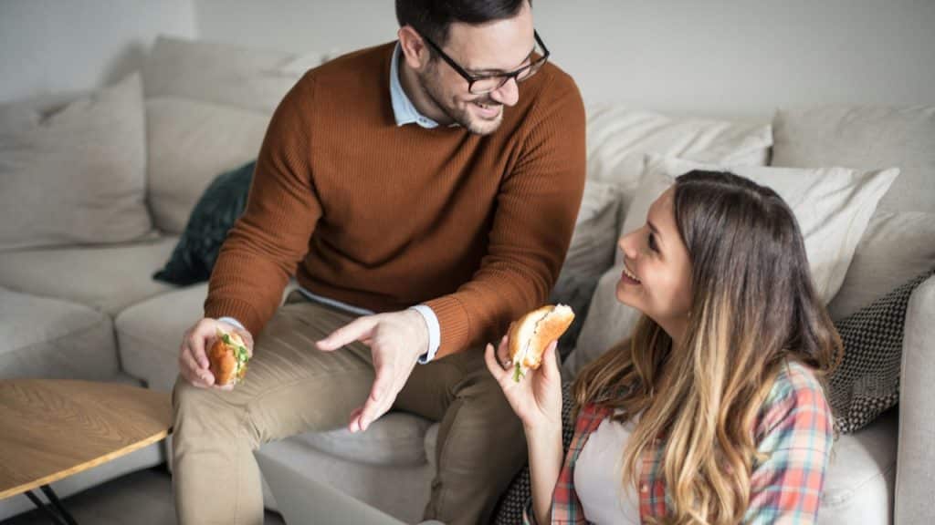 A smiling couple eats sandwiches while looking at a laptop in their living room.