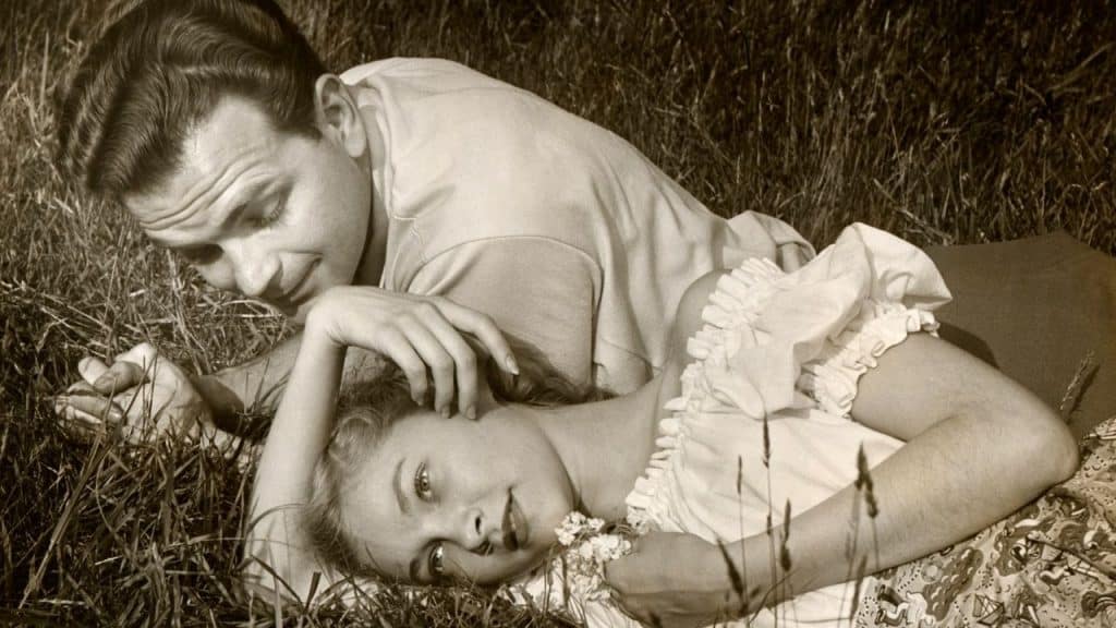 A sepia-toned photo of a man and woman lying in tall grass, with the man looking at the woman, and the woman looking at the camera.