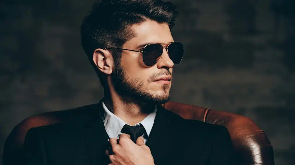 A man wearing sunglasses and a black suit is adusting his tie.