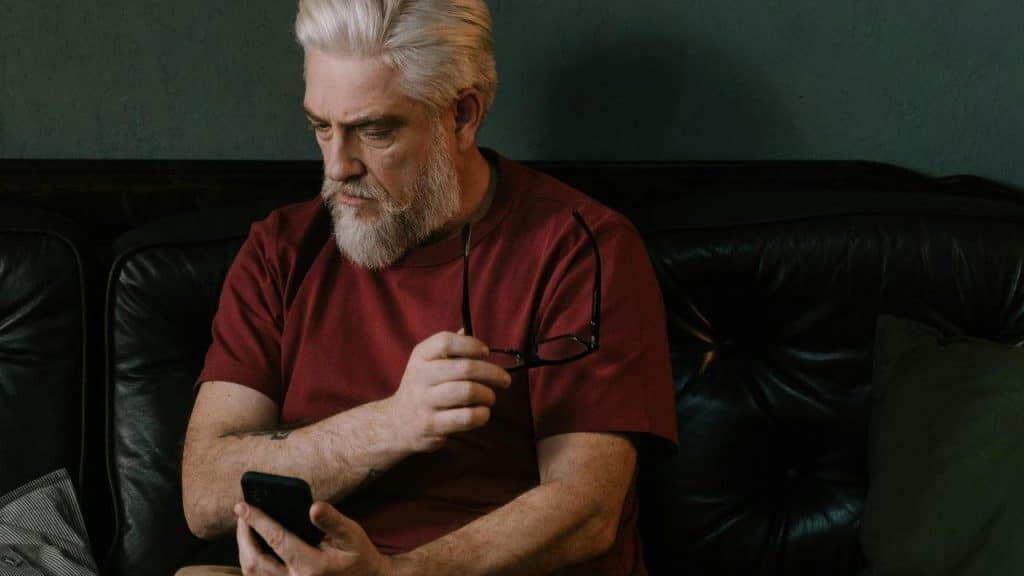 A man sitting on a couch looking at his phone while holding his glasses.