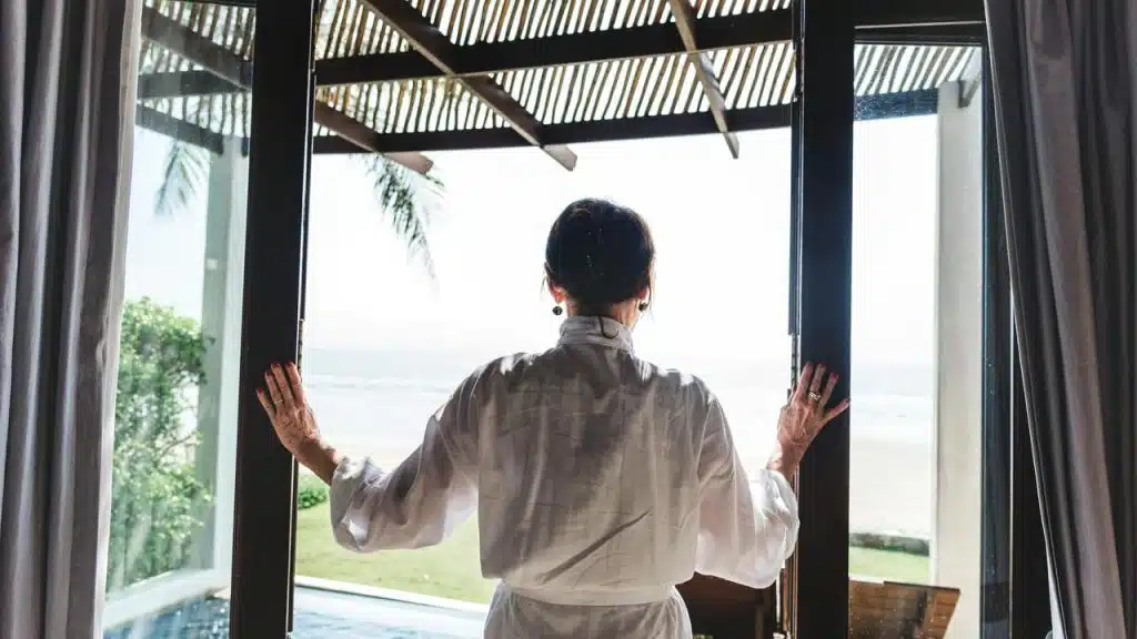 A woman in a robe opens glass doors to a bright beach view.