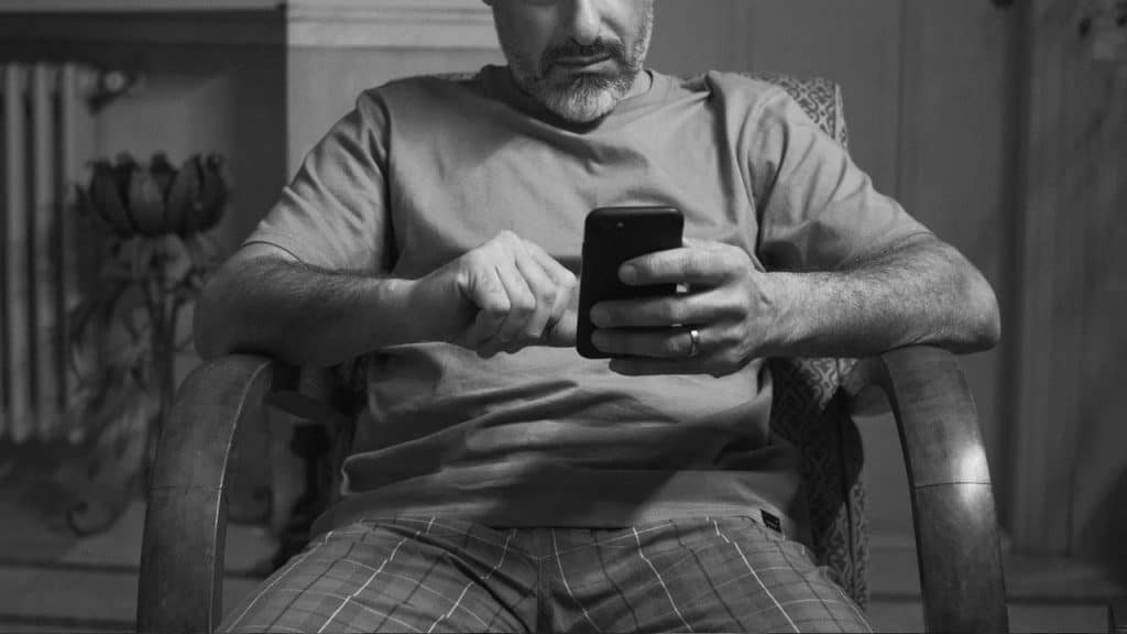Black and white close-up of a bearded man sitting in a wooden armchair looking down and using his smartphone.