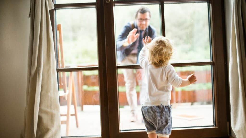 A blonde toddler looks out a glass door, with an adult waving back from outside.