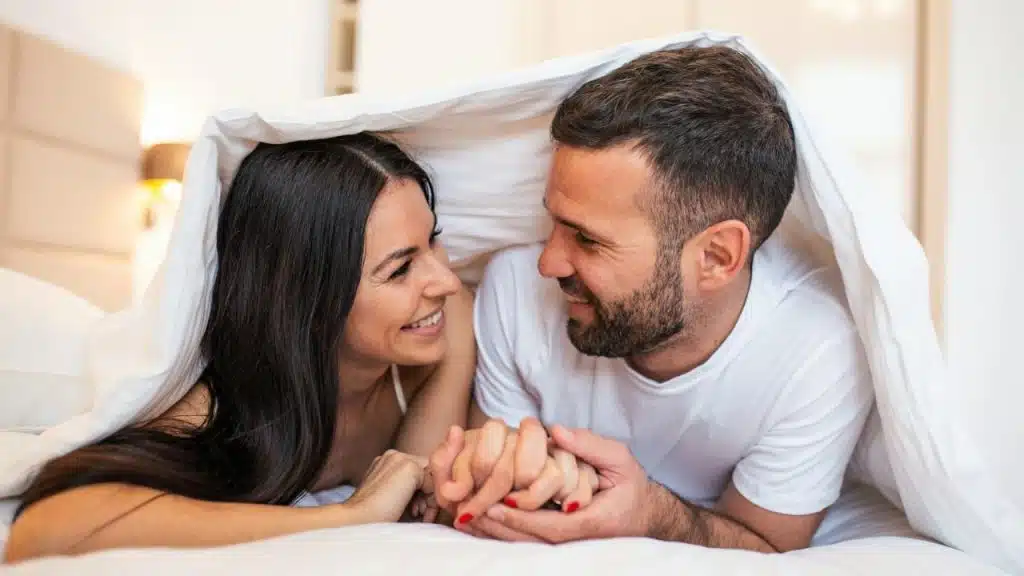 A smiling man and woman holding hands under a blanket in a bright bed.