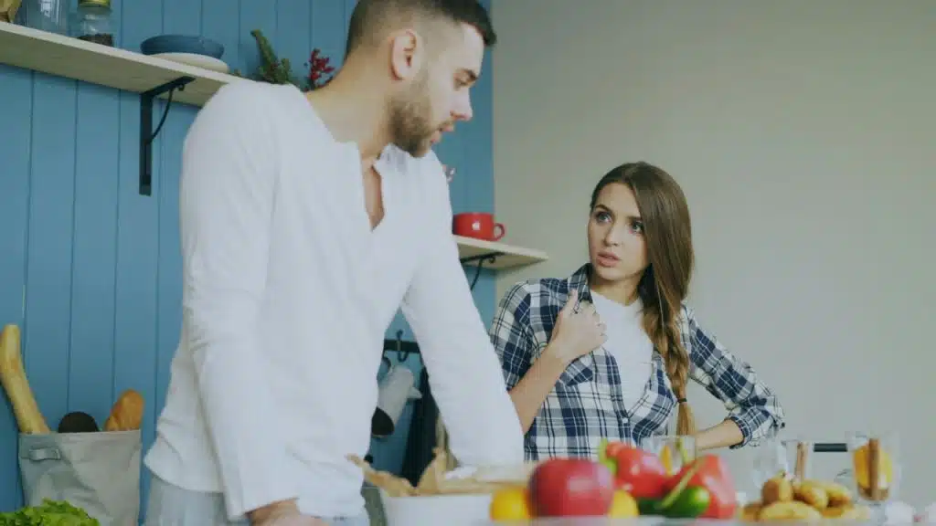 A man in a white shirt leans over a kitchen counter while a woman with a braid speaks to him.