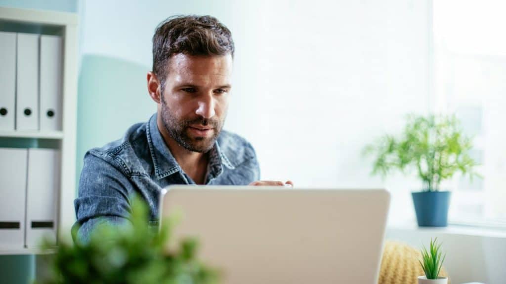 A man in a denim shirt is focused on working on a laptop at a desk.