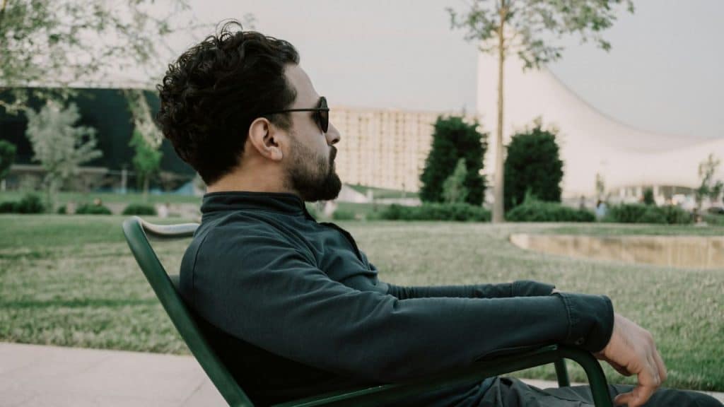 A man with a beard and sunglasses is sitting in a green chair outdoors, looking to the side.
