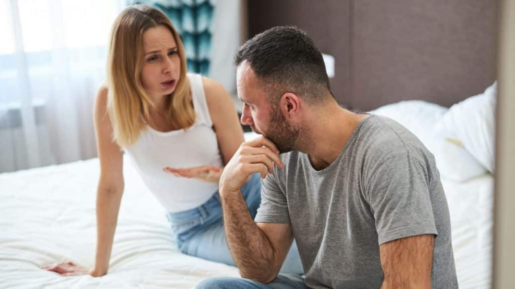 Woman actively talking to a man sitting on a bed with his hand resting on his chin.