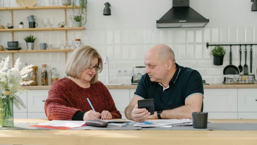 An older couple sits at a kitchen table, reviewing paperwork and holding a calculator.