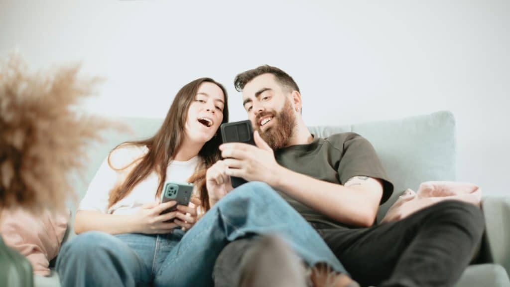 A smiling couple sits on a couch, both holding smartphones and looking at the man's screen.