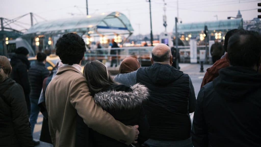 A crowd of people, including a couple with the man's arm around the woman, look toward a brightly lit, blurred urban background at dusk.