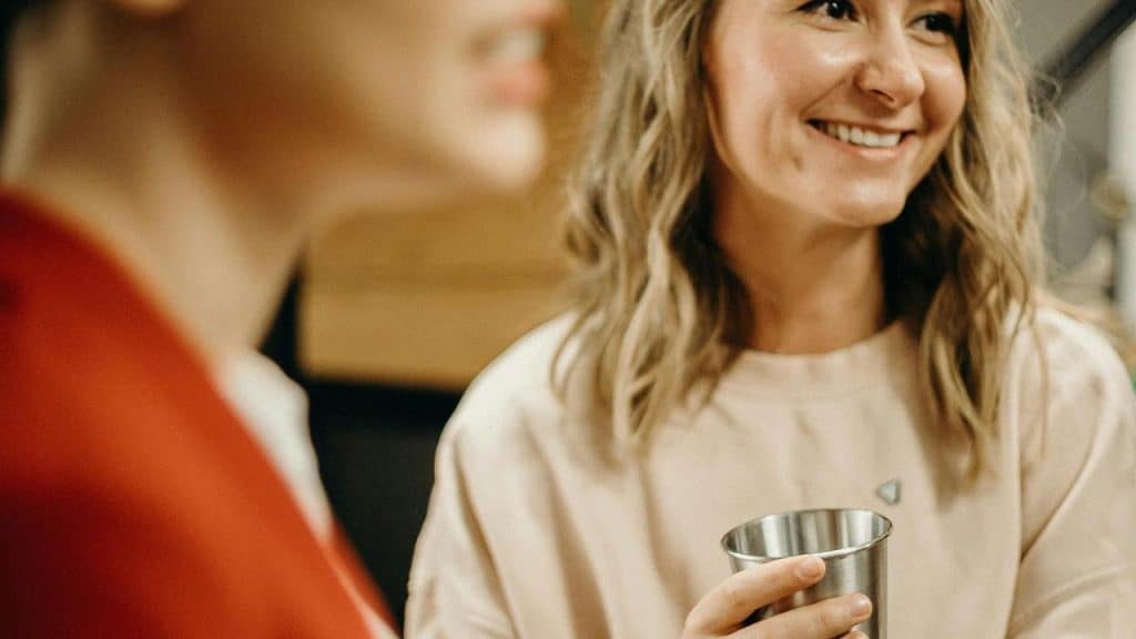 A smiling woman holding a metal cup while chatting with a friend.