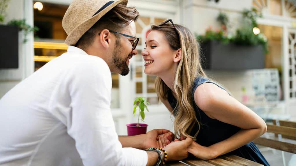 A couple leaning in and smiling while holding hands at an outdoor café.