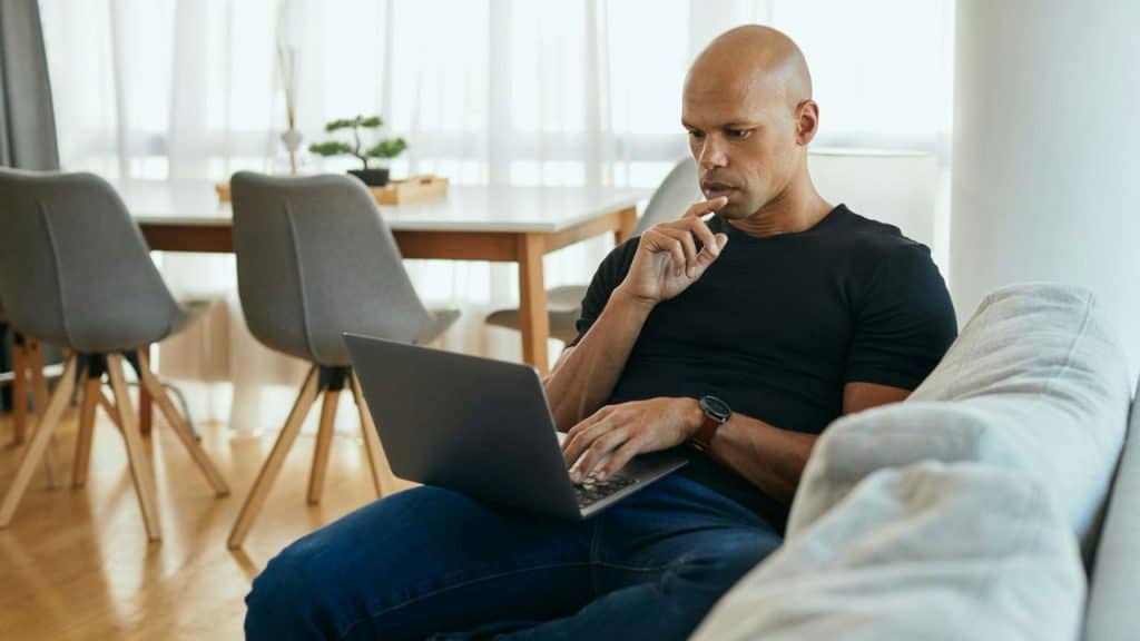 A bald man is sitting on a couch, working on a laptop with a thoughtful expression.