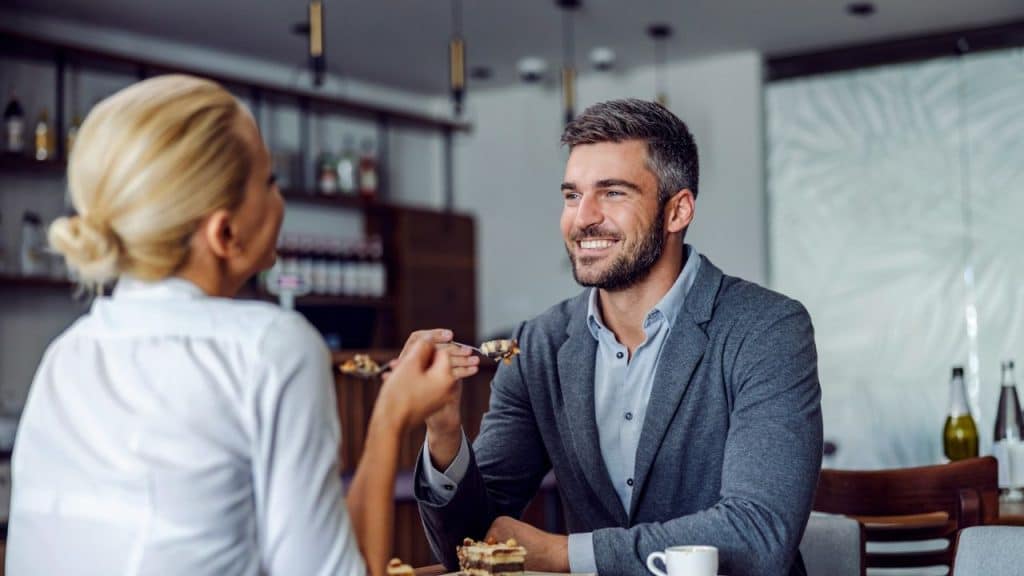 A man smiling while sitting across from a woman, being offered food on a spoon.