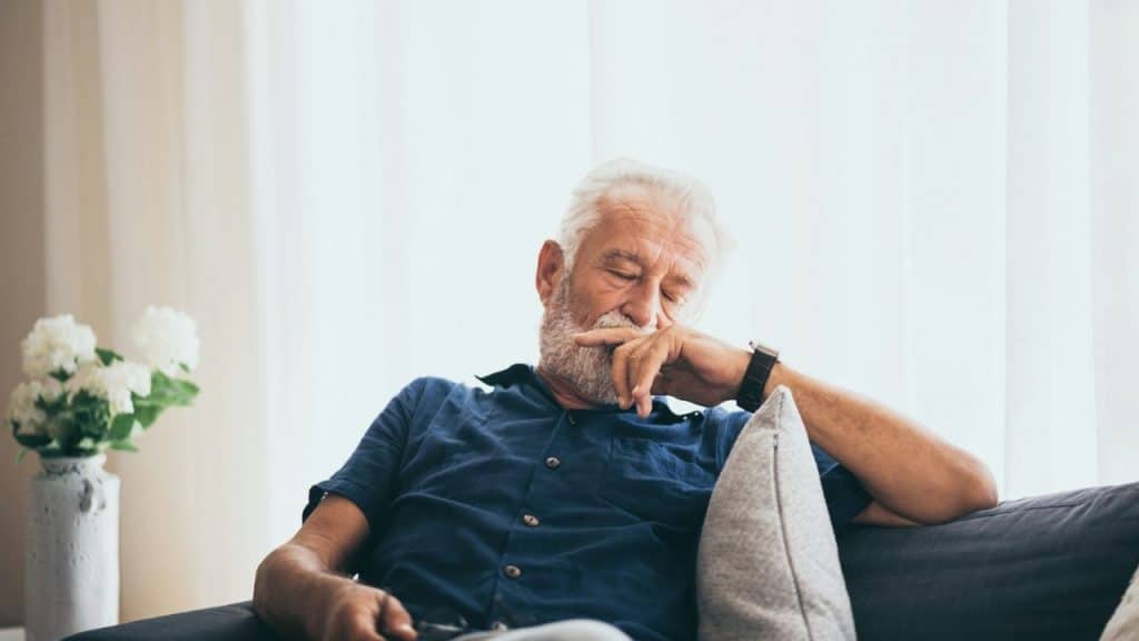 An older man with a white beard sits on a couch, holding his hand to his mouth.