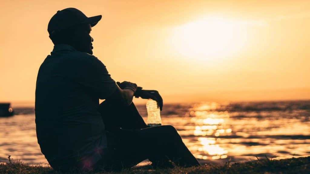 A silhouetted person wearing a baseball cap sits by the water, holding a plastic bottle at sunset.