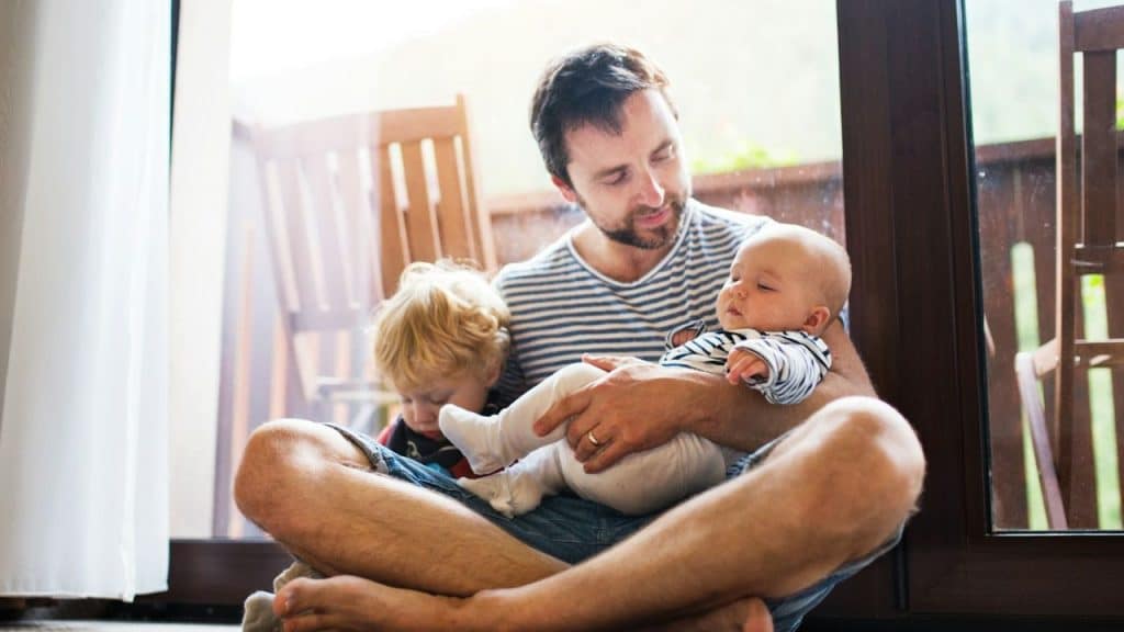 A father sits cross-legged, holding a sleeping baby and a toddler.