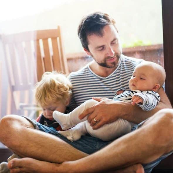 A father sits cross-legged, holding a sleeping baby and a toddler.