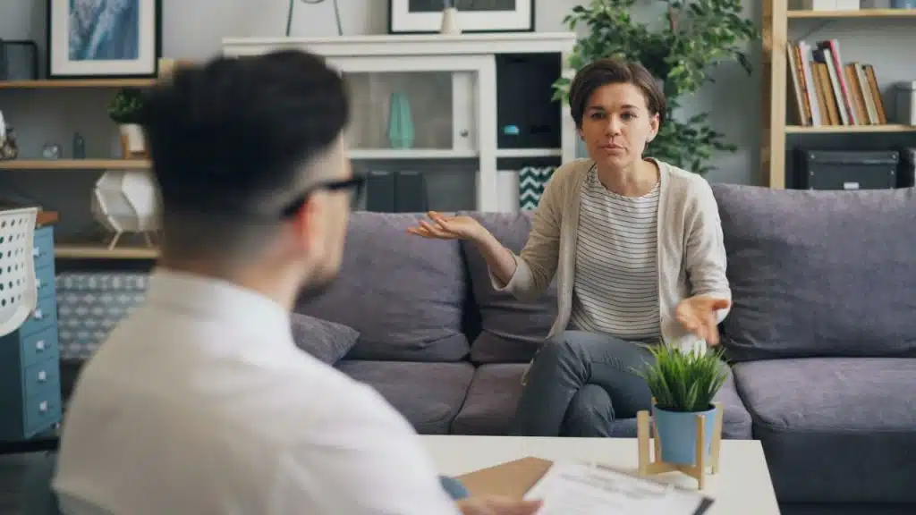 Woman on a gray couch talking to a man with her hands open, in a counseling or discussion setting.