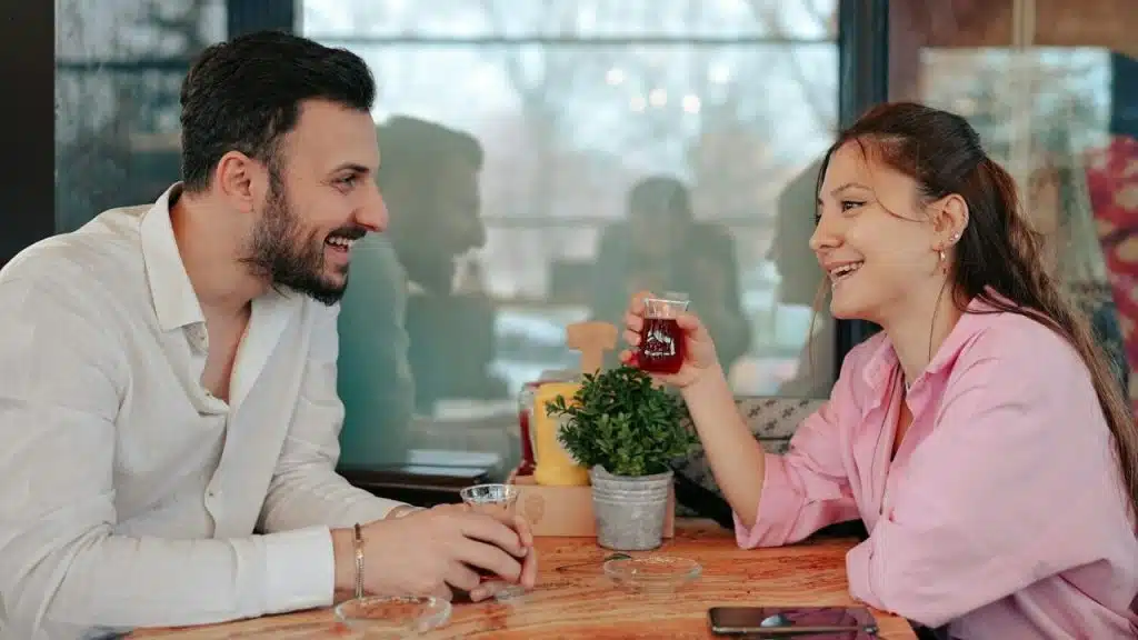 Smiling man and woman seated at a table, drinking from small clear glasses.