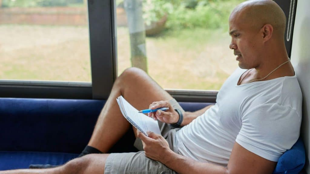Bald man in a white t-shirt and shorts sitting on a blue couch by a window, writing in a notebook.