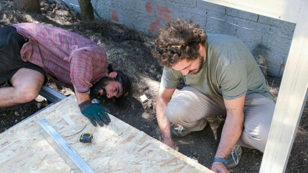 Two bearded men are working together on a wooden panel outdoors.