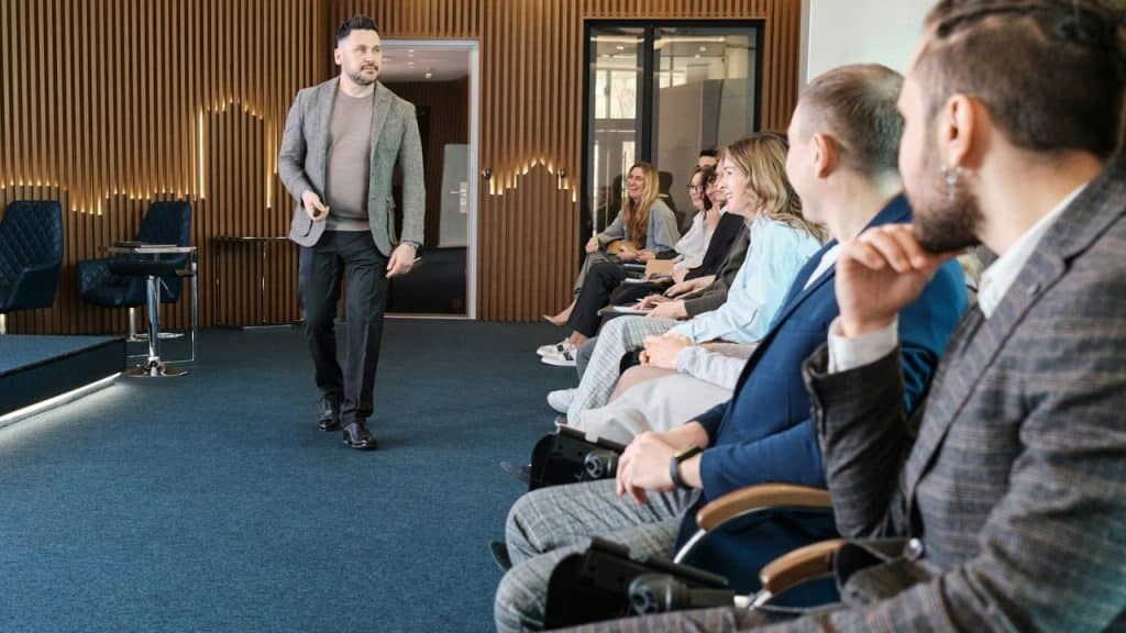 A speaker in a tweed jacket is walking toward an audience in a modern room.