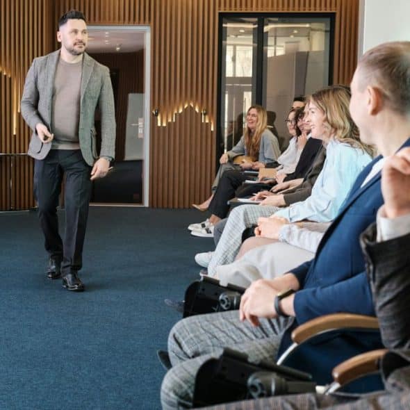 A speaker in a tweed jacket is walking toward an audience in a modern room.