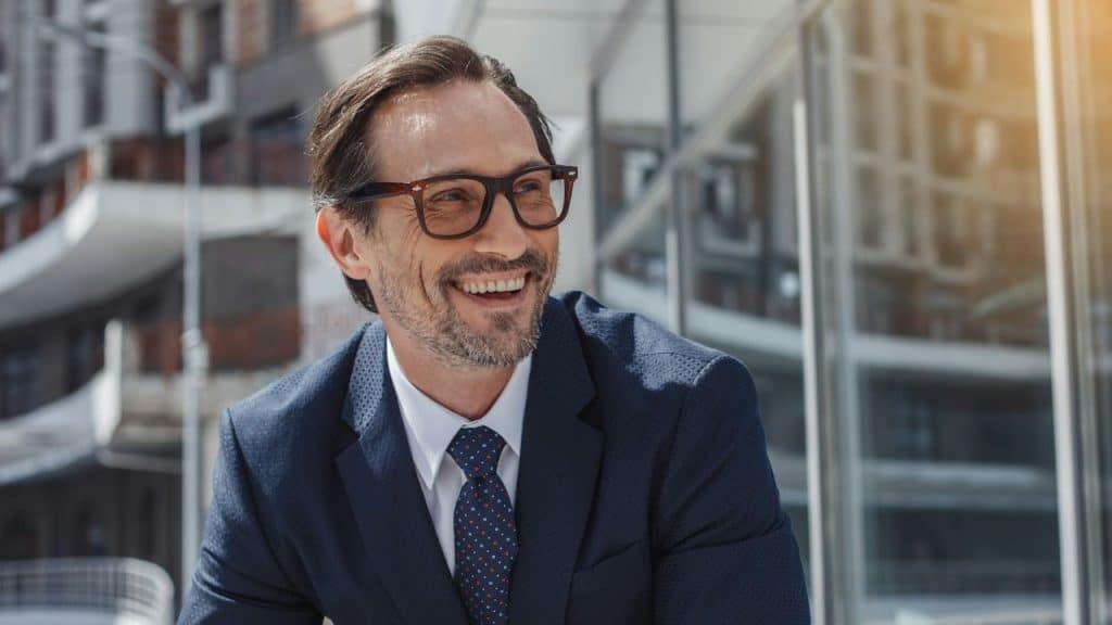 Smiling, middle-aged man in a navy suit and glasses stands outside a modern building.