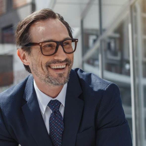 Smiling, middle-aged man in a navy suit and glasses stands outside a modern building.