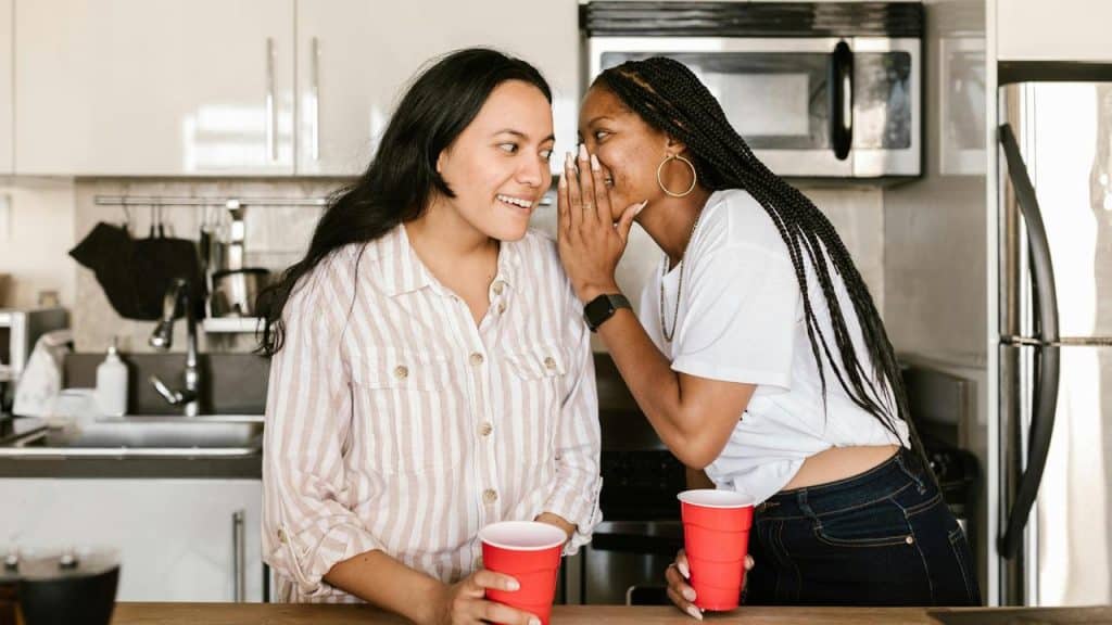 A woman whispering to another woman while they hold red cups in a kitchen.