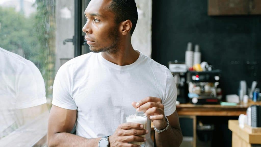 A man in a white t-shirt holding a drink while looking out the window.