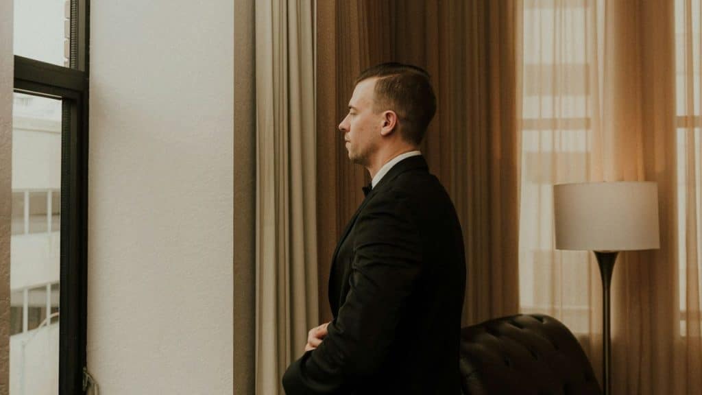 Man in a tuxedo standing in profile by a window in a well-lit room.