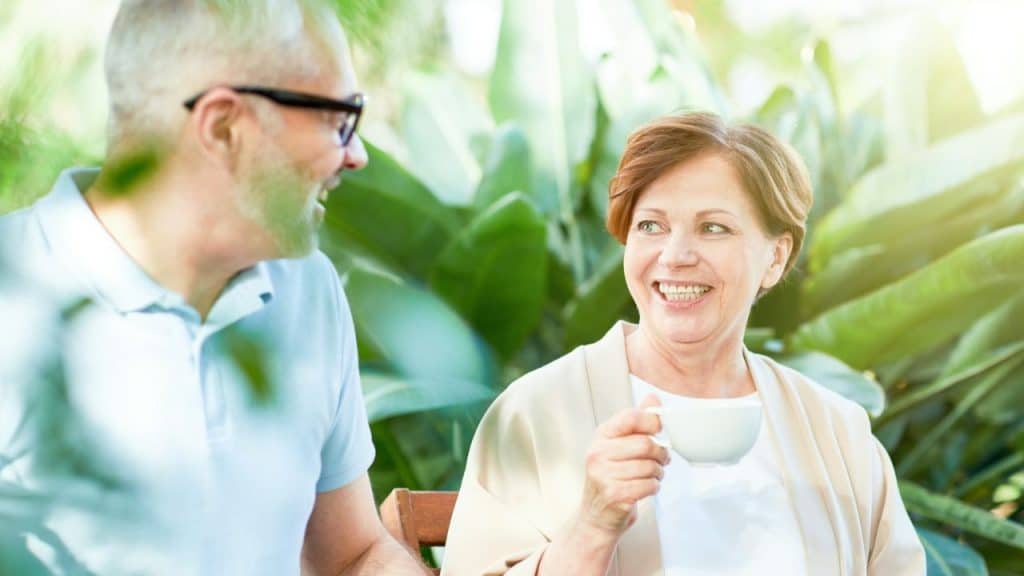 Smiling woman holding a coffee cup looking at a man, surrounded by lush green plants.