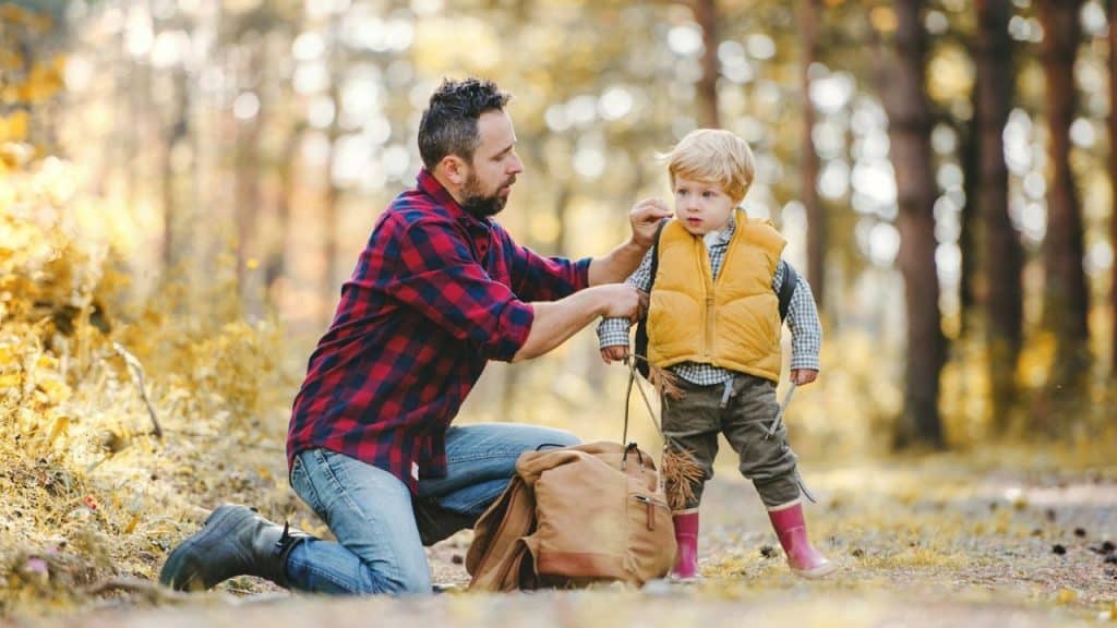 Man kneels next to a backpack in a forest, adjusting a boy's yellow vest.
