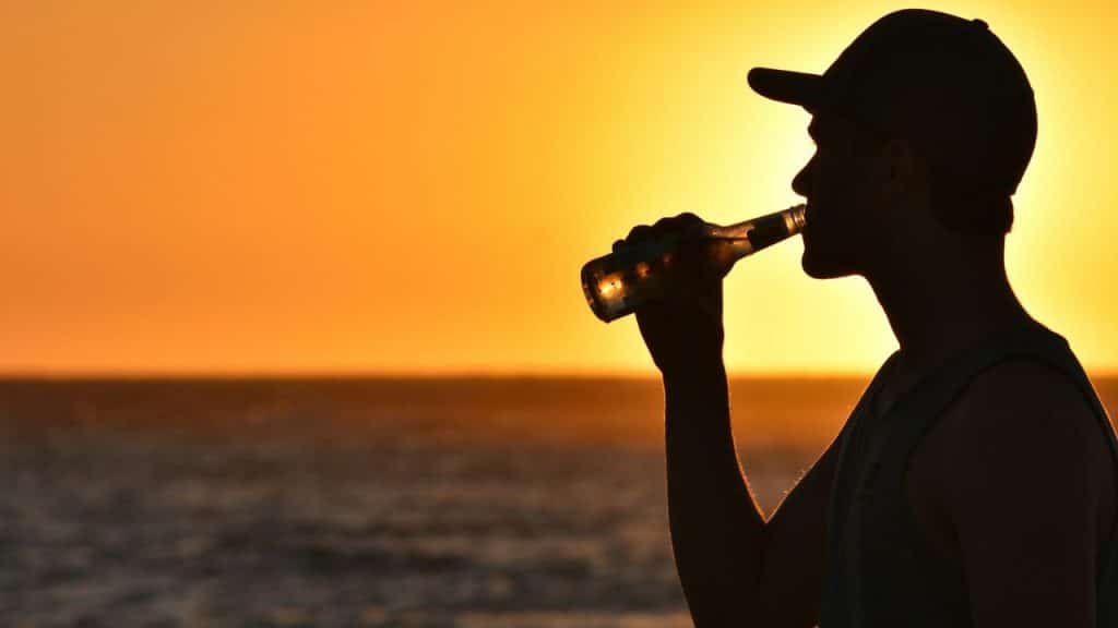 Silhouette of a person in a baseball cap drinking from a bottle at sunset by water.
