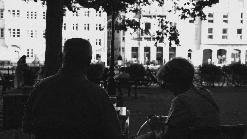 A high-contrast, black and white image showing the dark silhouettes of two people sitting at an outdoor table with brightly lit buildings in the background.