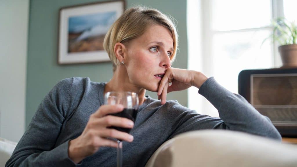 A blonde woman on a couch holds a glass of red wine, looking away with a pensive expression.