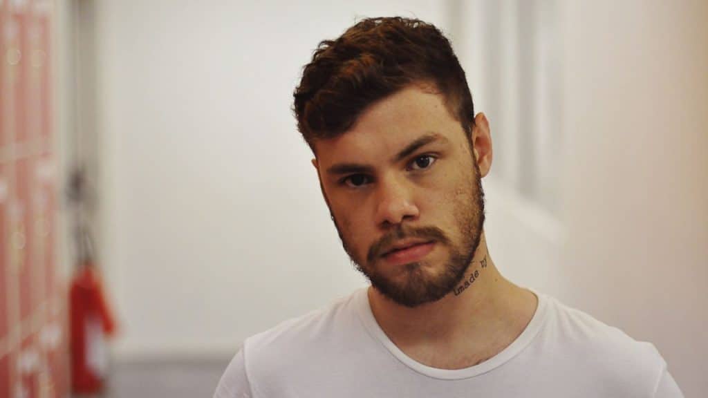 A young man with a beard and a neck tattoo looks directly at the camera in a white hallway.