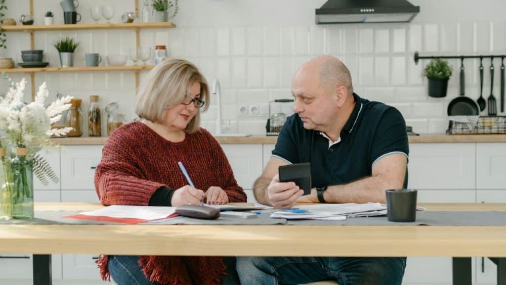 An adult couple sits at a wooden table in a kitchen. The woman is writing on papers, and the man holds a calculator..