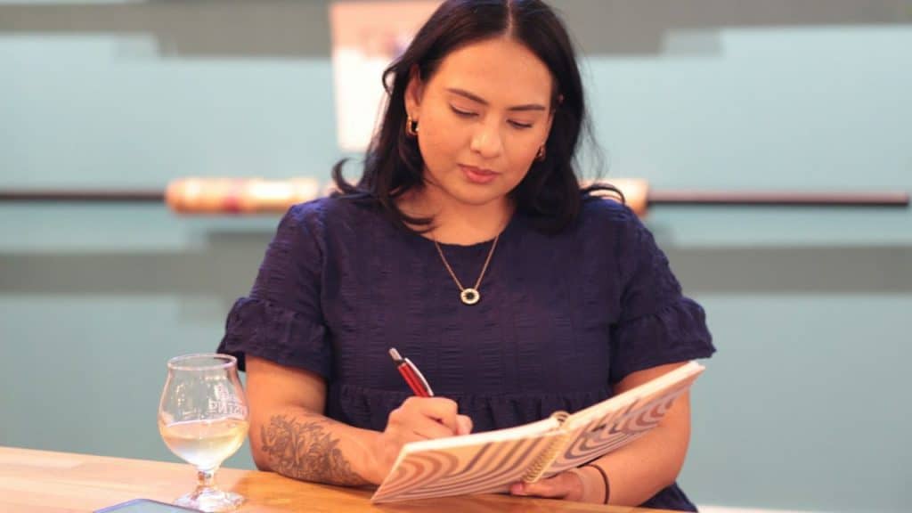 A woman in a dark blue dress writes in a notebook beside a glass of white wine.