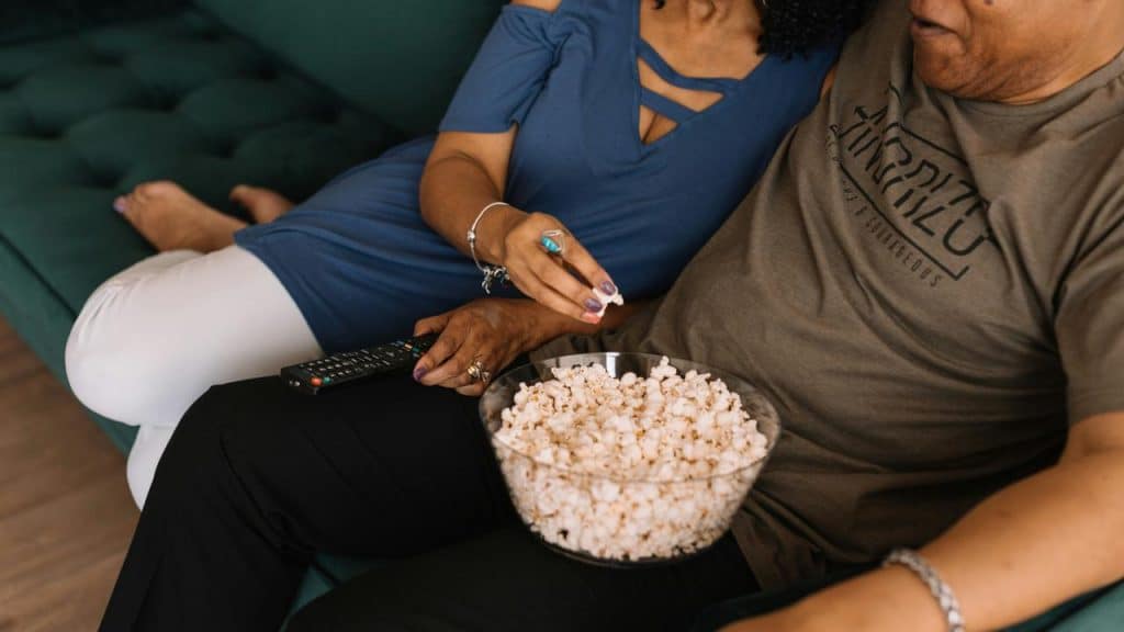 A couple sitting on a couch sharing a bowl of popcorn while watching TV.