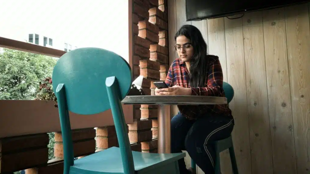 Woman in glasses and plaid shirt sitting at a table, looking at her phone.