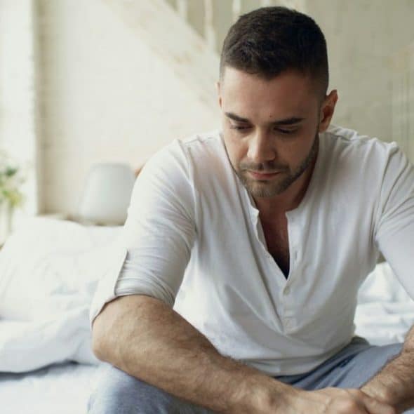 Man in white shirt sitting on bed, looking down, with person behind him.