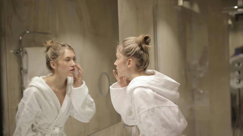 A woman in a bathrobe looking at herself in the bathroom mirror.
